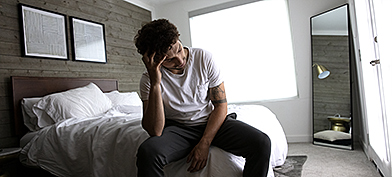 A man sits on the bed holding his head in his hand.