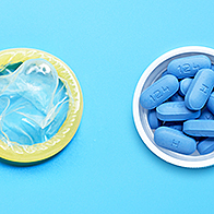 Blue pills sit in a small open white container next to a condom on a blue surface.