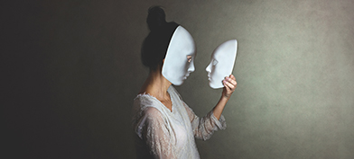 A woman in a white mask holds another mask in her hand and looks at it face to face.