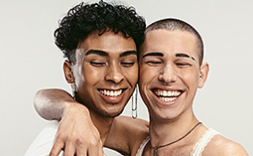 One person hugs their right arm around another person as both smile with their eyes closed.