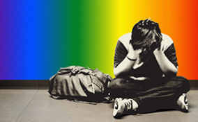A child sits on the ground with his legs crossed and head in his hands against a rainbow wall.