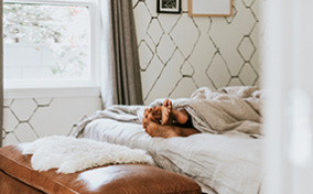 The feet of a couple shows from under the blankets while they lie in bed together.