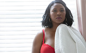 A woman in a red bra holds a white blanket and stands in front of a window with drawn blinds.